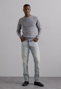 Maglione grigio a maglia, jeans azzurri chiari con sbiaditura e scarpe nere. Il modello è in piedi con le mani in tasca su uno sfondo neutro.
