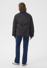 Černá puffer bunda s vysokým límcem, zdobená chevronovým švem a volným střihem, v kombinaci s modrými džíny a bílou obuví.