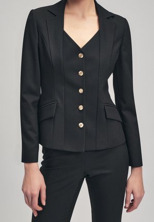 Woman wearing a fitted black blazer with five gold buttons, flap pockets, and matching black pants against a neutral background.