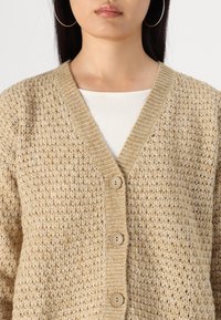 Beige strikket cardigan med V-hals, med et teksturert mønster, fem knapper og ribbestrikket kant langs kragen og fronten.