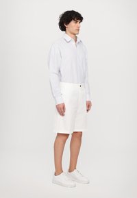 Hvite bomullsshorts med oppbrettede hem, sammen med en lyseblå rutete skjorte med lange ermer. Brukt med hvite sneakers.