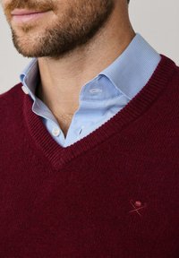 Pull en V bourgogne en tricot doux, porté par-dessus une chemise à carreaux bleu clair. Présente un petit logo brodé sur le pull.