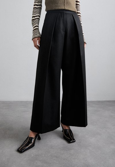 Pantalons noirs à jambes larges en tissu texturé, avec des plis, une taille haute, et accompagnés de chaussures à talons noires.