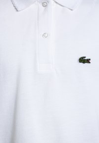 Biała koszulka polo wykonana z teksturowanej tkaniny, z dwoma guzikami przy dekolcie i zielonym haftowanym logo krokodyla na lewej piersi.