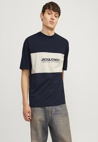 Marineblå og cremefarvet kortærmet t-shirt med rund hals. Har brandlogo på brystet. Sammen med tilfældigt slidte denimjeans. Blødt bomuldsstof.