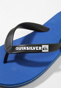 Tongs bleues avec une semelle texturée, une sangle en caoutchouc noir et le logo "Quiksilver" en blanc. Design plat et léger, adapté à une utilisation décontractée.