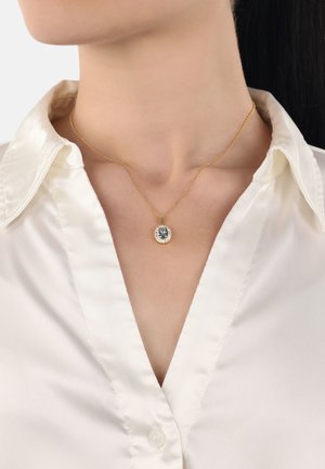 Collier en chaîne dorée avec un pendentif ovale, présentant une pierre centrale entourée de petites pierres claires, porté sur une chemise en satin blanche.