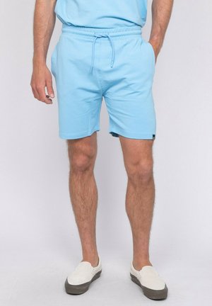 Homme portant un short bleu clair à cordon et un t-shirt assorti, debout les mains dans les poches, chaussures blanches sans lacets visibles.