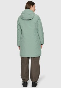 Femme portant un manteau à capuche vert sauge, un pantalon cargo marron et des bottes noires, se tenant de dos sur un fond neutre.