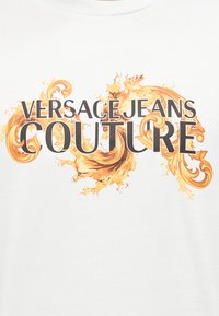 Άσπρο ύφασμα με το κείμενο «VERSACE JEANS COUTURE» που καλύπτει χρυσές διακοσμητικές σπείρες και σχεδιασμούς με πιτσιλιές.