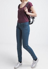 Bordeaux top met kantdetails, blauwe strakke broek en zilveren sneakers; draagt een zwarte en zilveren rugzak. Zachte stoffen, modern ontwerp.
