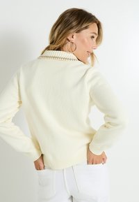 Suéter de punto color crema con cuello acanalado y acentos de cuentas, mangas largas y un diseño ajustado, combinado con pantalones blancos.