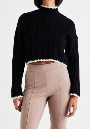 Kvinde iført sort ribbet cropped sweater med hvid kant og højtaljede beige tætsiddende bukser mod en ensfarvet baggrund.