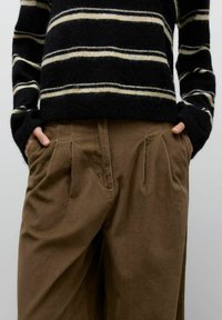 Person trägt braune, gefaltete Cordhosen, hat die Hände in den Taschen und einen schwarzen Pullover mit beigen horizontalen Streifen.