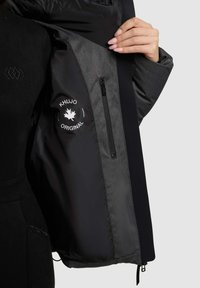 Sort jakke med grå indvendig foring, med en lynlåslomme og et hvidt logo patch. Glat tekstur og moderne design detaljer.