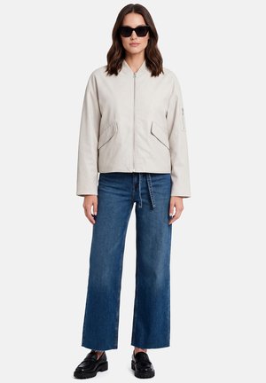 Frau trägt eine hellbeige Reißverschlussjacke, weite blaue Jeans mit gebundenem Gürtel, schwarze Loafers und schwarze Sonnenbrille, steht vor einem weißen Hintergrund.