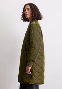 Olijfgroene, doorstikte lange jas met ribbelmanchetten, zijzakken en een ritszak op de mouw; gladde, gewatteerde textuur met een relaxte pasvorm.