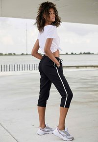Zwarte sportieve cropped broek met witte zijkanten, gecombineerd met een vastgebonden wit T-shirt. Heeft krullend haar en witte sneakers met roze accenten.