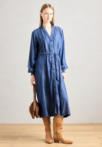 Denimska obleka z gumbi spredaj, gubanimi manšeti in vezanim pasom. V kombinaciji z rjavimi čevlji do kolen in rjavim ročnim torbico.