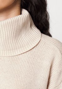 Beżowy sweter z golfem w prążki noszony przez osobę z długimi ciemnymi włosami, z widocznymi detalami szyi i ramion na neutralnym tle.