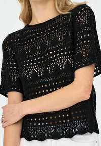 Žena oblečená v černém krátkém rukávu háčkovaném topu s geometrickými vzory a vroubkovaným lemem, spárovaná s bílými kalhotami, ruka zkřížená na pase.