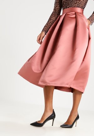 Vrouw die een roze satijnen midi-rok draagt, een langarmige top met luipaardprint en zwarte pumps met hoge hakken en een spits toelopende neus, staand tegen een witte achtergrond.