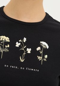 Camiseta de algodón negra con diseños florales en dorado metálico y blanco, con el texto "sin lluvia, no hay flores" debajo de los gráficos.