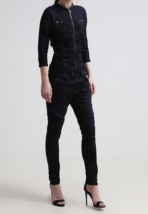 Nainen, joka on pukeutunut istuvaan mustaan vetoketjulliseen takkiin ja yhteensopiviin kapeisiin housuihin, seisoo mustissa avoimilla varpailla varustetuissa korkokengissä yksiväristä taustaa vasten.