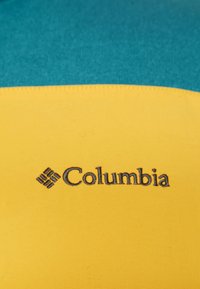 Logo Columbia su un tessuto con strisce orizzontali turchesi e gialle, texture liscia e dettagli di cucitura.