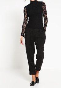 Top in pizzo nero a maniche lunghe con collo alto; abbinato a pantaloni neri larghi. I pantaloni presentano una vestibilità ampia e un dettaglio a pieghe sui polsini.