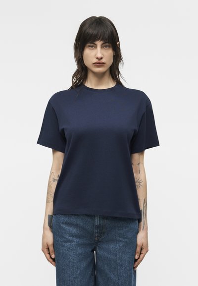 Kvinde med mørkt hår iført en navyblå T-shirt med korte ærmer og blå jeans, stående foran en ensfarvet lys baggrund.