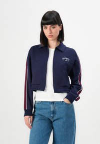 Navy cropped sweatshirt with a collar, zip closure, and "GAP NYC" logo. Features maroon side stripes and ribbed cuffs, paired with blue jeans.