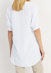 Blusa bianca con una vestibilità rilassata, maniche corte a sbuffo e dettagli smock ai polsini. Il tessuto sembra leggero e liscio.