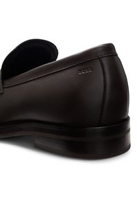 Mocasín de cuero marrón oscuro con una textura suave, suela plana y tacón de madera apilada. Cuenta con el logo de la marca en relieve en el lateral.