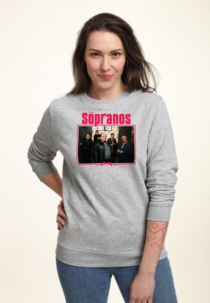Femme aux longs cheveux bruns portant un sweat-shirt gris avec la photo et le logo du casting de The Sopranos, debout avec une main sur la hanche, fond neutre.