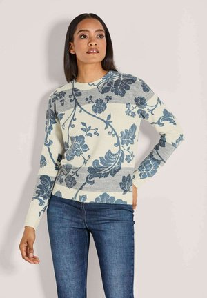 Frau in einem cremefarbenen Pullover mit blauen Blumenmustern und grauen Streifen, kombiniert mit dunkelblauen Jeans, steht vor einer hellen Wand.