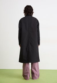 Personne avec des cheveux bouclés portant un long manteau noir et un pantalon mauve à jambes larges, se tenant sur un sol vert devant un mur blanc, dos à la caméra.