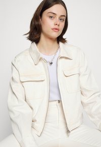 Jaqueta de ganga off-white com colarinho, duas bolsos no peito e fecho éclair na frente. Usada sobre uma blusa branca e combinada com calças de cor clara.