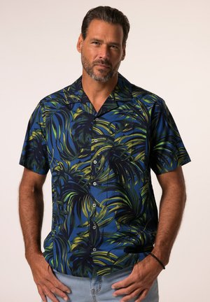 Man met baard draagt een blauw kort mouw overhemd met knopen en groene en zwarte tropische bladpatronen, staand met handen bij de zakken.