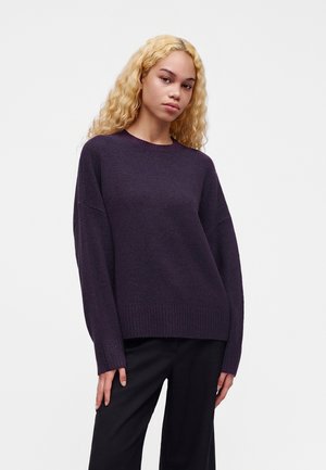 Giovane donna con lunghi capelli biondi ricci che indossa un maglione viola scuro e pantaloni neri, in piedi davanti a uno sfondo chiaro e uniforme.