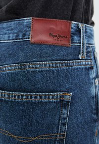 Modri džins hlače z rjavim usnjenim žetonom z napisom "Pepe Jeans London", kontrastni šivi in zadnjimi žepi.