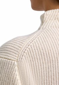 Maglione lavorato a maglia beige chiaro con collo alto, caratterizzato da costine testurizzate e cuciture delle spalle evidenti. Il filato morbido e spesso crea un aspetto accogliente.