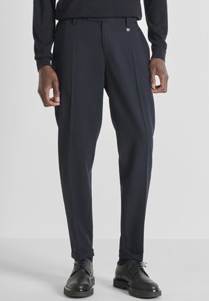 Pantalones pantalón de marino con pliegues, confeccionados en una tela suave con una cintura ajustada, que presenta un pequeño logo. Combinados con zapatos negros de cordones.