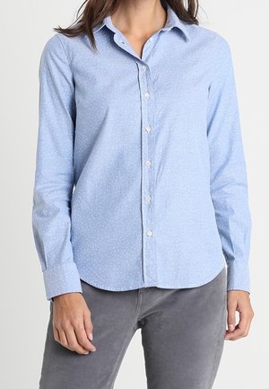 Jasnoniebieska koszula z długim rękawem zapinana na guziki z małymi białymi kropkami, noszona z szarymi spodniami, pokazana od szyi do bioder.