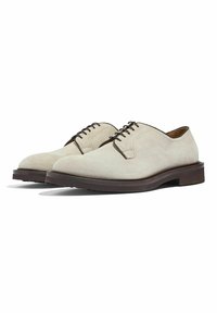 Scarpe in suede beige con una texture liscia, caratterizzate da una punta tonda, lacci marroni e una suola in gomma marrone scuro. Design minimalista con linee pulite.