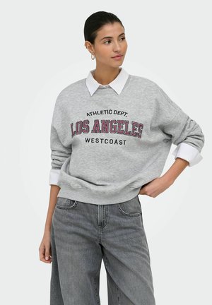 Kvinde med mørkt hår iført en grå sweatshirt med teksten Los Angeles Westcoast over en hvid skjortekrave og løse grå jeans, poserer med den ene hånd på hoften.