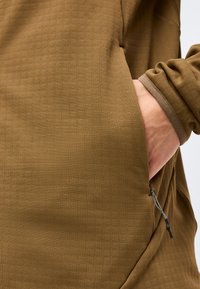 Το χέρι ενός ατόμου έχει τοποθετηθεί στην πλαϊνή τσέπη ενός καφέ μακρυμάνικου μπουφάν με ορατό φερμουάρ στην τσέπη.