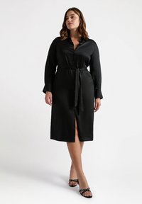 Vestito nero con bottoni, maniche lunghe e cintura in vita, con uno spacco frontale. Texture liscia e silhouette aderente. Indossato con sandali neri.