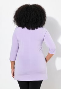 Top lavanda chiaro con maniche a tre quarti, texture liscia, parte posteriore più lunga, orlo arrotondato, indossato sopra leggings neri, visto da dietro.