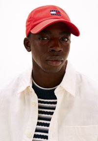 Rød cap med logo-patch, båret med en hvid skjorte og en navy stribet sweater. Nærbillede, der fremhæver ansigt og tøj.